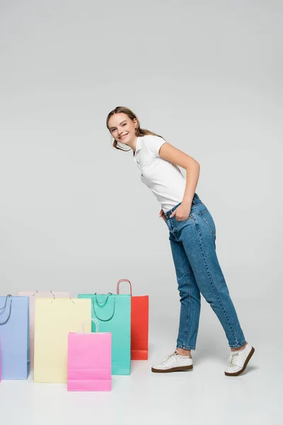 Mujer alegre de pie con las manos en los bolsillos cerca de bolsas de compras en gris, concepto de viernes negro - foto de stock