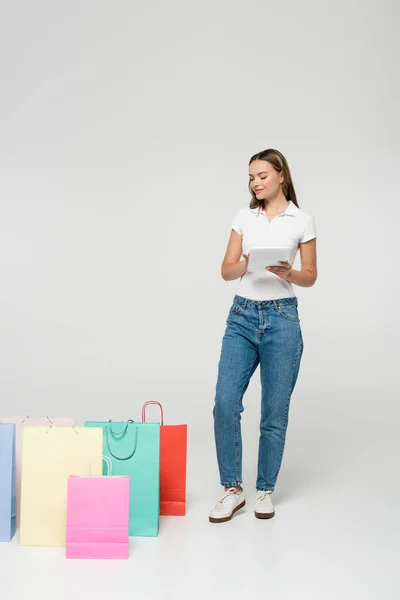 Joyful woman standing and holding digital tablet near shopping bags on grey, black friday concept — Stock Photo