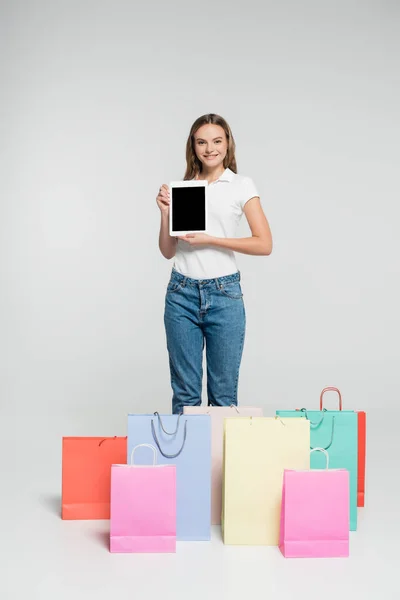 Freudige Frau steht und hält digitales Tablet mit leerem Bildschirm neben Einkaufstaschen am grauen, schwarzen Freitag — Stockfoto