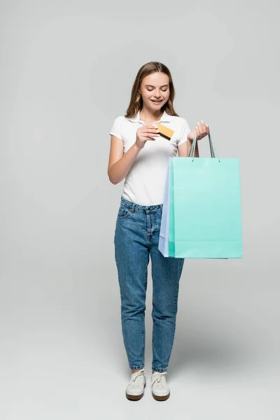 Alegre joven mujer sosteniendo tarjeta de crédito y mirando azul bolsas de compras en gris, concepto de viernes negro - foto de stock