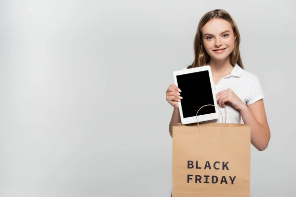 Alegre joven mujer sosteniendo tableta digital con pantalla en blanco cerca de la bolsa de compras con letras de viernes negro en gris - foto de stock