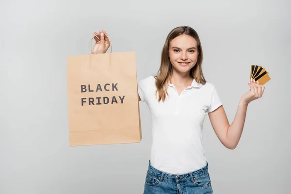 Pleased woman holding credit cards and shopping bag with black friday lettering isolated on grey — Stock Photo
