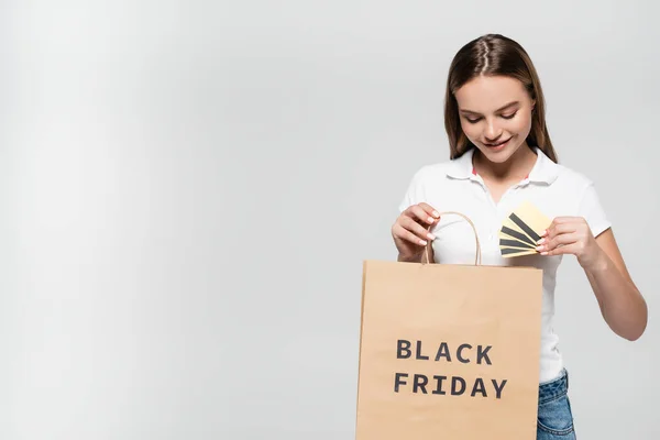 Mulher alegre segurando cartões de crédito e olhando para o saco de compras com letras sexta-feira preto isolado em cinza — Fotografia de Stock
