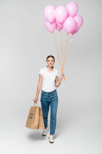 Young joyful woman holding shopping bags with black friday lettering and pink balloons on grey — Stock Photo