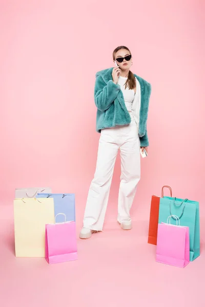 Femme élégante en lunettes de soleil parler sur smartphone près des sacs à provisions sur rose, concept vendredi noir — Photo de stock