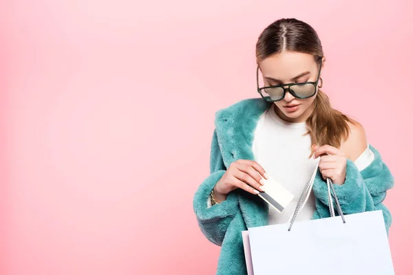 Молода модна жінка в сонцезахисних окулярах тримає кредитну картку біля сумки для покупок ізольована на концепції рожевої, чорної п'ятниці — стокове фото