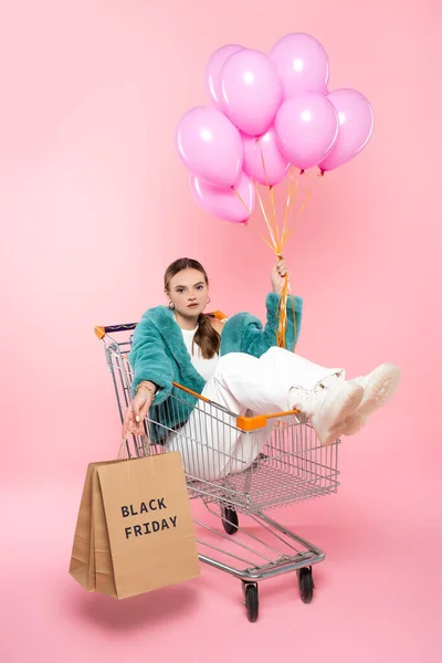 Young woman sitting in cart with black friday lettering on shopping bags and holding balloons on pink — Stock Photo