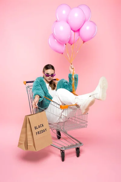 Young woman in sunglasses sitting in cart with black friday lettering on shopping bags and holding balloons on pink — Stock Photo
