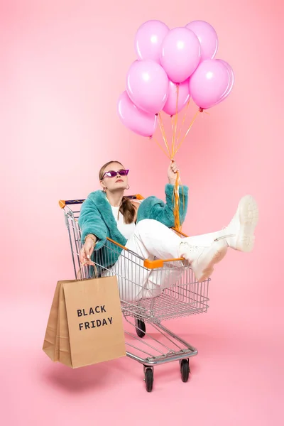 Стильна жінка в сонцезахисних окулярах, що сидить у візку з чорною п'ятничною пляшкою на сумках для покупок і тримає повітряні кулі на рожевому — стокове фото