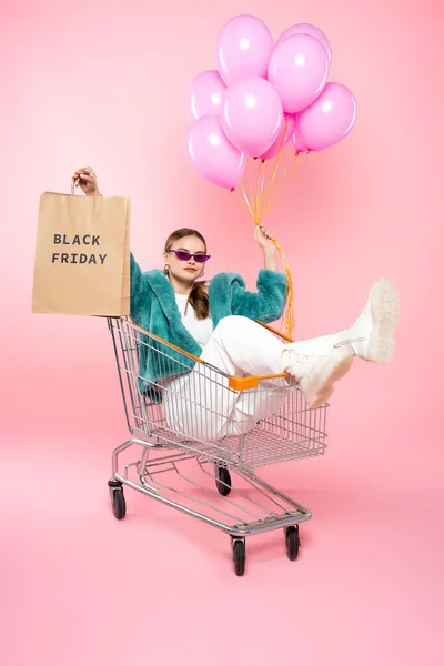 Stylish woman in sunglasses sitting in cart with black friday lettering on shopping bag and holding balloons on pink — Stock Photo