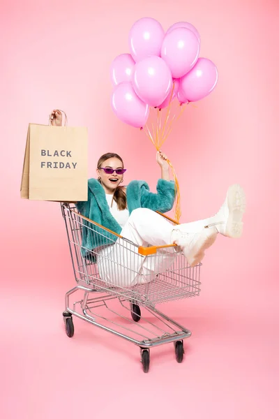 Femme à la mode dans des lunettes de soleil assis dans le panier avec lettrage noir vendredi sur le sac à provisions et tenant des ballons sur rose — Photo de stock