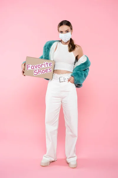 Mujer joven en la caja de la máscara médica con las letras favoritas de los zapatos mientras está de pie con la mano en el bolsillo en rosa — Stock Photo