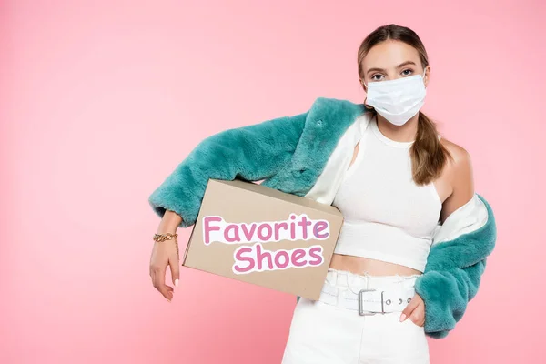 Junge Frau in medizinischer Maske hält Box mit Lieblingsschuhen Schriftzug auf rosa — Stockfoto