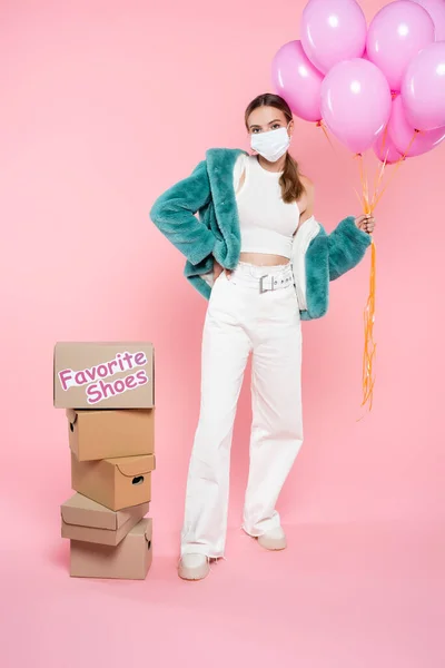 Молодая женщина в медицинской маске держит воздушные шары возле коробки с любимыми туфлями, написанными на розовом — стоковое фото
