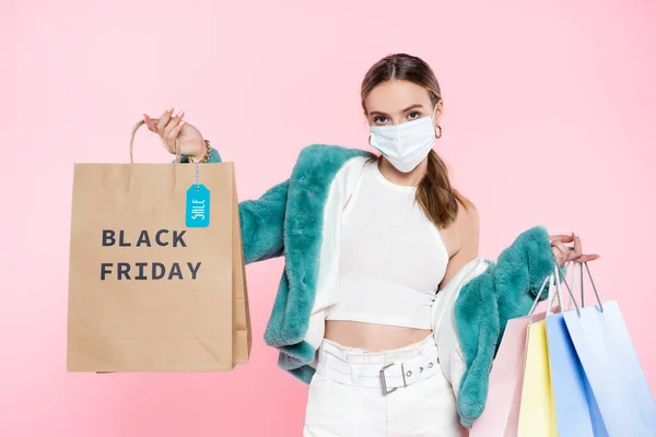Молодий клієнт в медичній масці тримає сумку для покупок з чорною п'ятницею з написом та тегом продажу ізольовано на рожевому — стокове фото