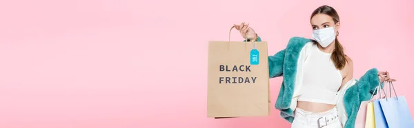 Panoramic shot of young customer in medical mask holding shopping bag with black friday lettering and sale tag isolated on pink — Stock Photo