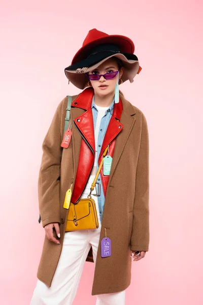 Jeune femme en lunettes de soleil et chapeaux avec des étiquettes de vente debout sur rose — Photo de stock