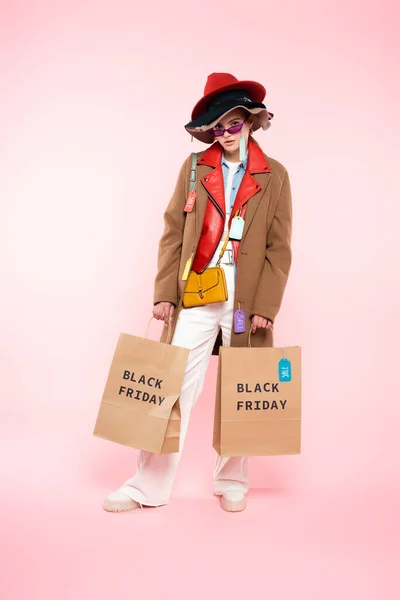 Jeune femme en lunettes de soleil et chapeaux avec des étiquettes de vente debout avec des sacs à provisions sur rose, concept vendredi noir — Photo de stock