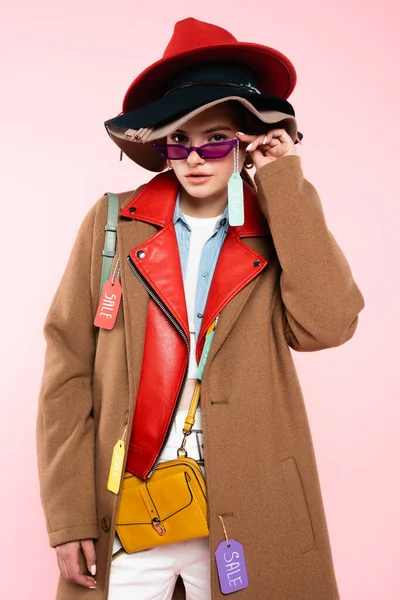 Jeune femme à la mode dans des chapeaux touchant lunettes de soleil avec étiquette de vente sur rose — Photo de stock