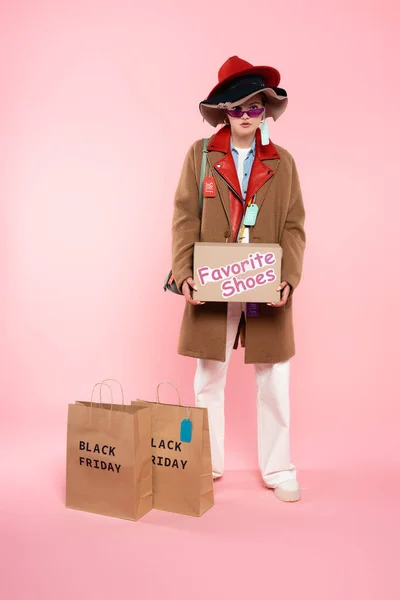 Femme en lunettes de soleil et chapeaux avec des étiquettes de vente tenant boîte en carton avec des chaussures préférées lettrage près de sacs à provisions sur rose, concept vendredi noir — Photo de stock