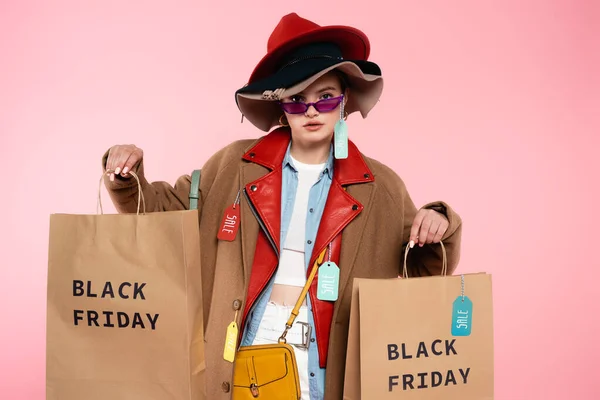 Trendige Frau in Sonnenbrille und Hut mit Verkaufsetiketten, die Einkaufstaschen auf rosa, schwarzem Freitag-Konzept halten — Stockfoto