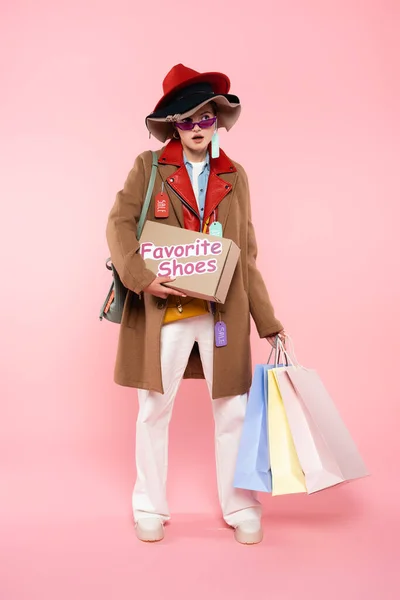 Femme surprise dans des lunettes de soleil et des chapeaux avec des étiquettes de vente tenant boîte avec chaussures préférées lettrage et sacs à provisions sur rose, concept vendredi noir — Photo de stock