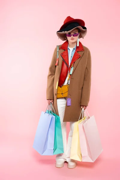 Mujer en gafas de sol y sombreros con etiquetas de venta sosteniendo bolsas de compras y de pie en rosa - foto de stock