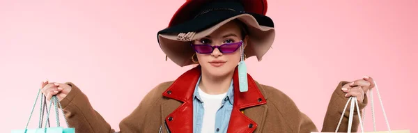 Tiro panorâmico de mulher em óculos de sol e chapéus com venda tag segurando sacos de compras isolados em rosa — Fotografia de Stock