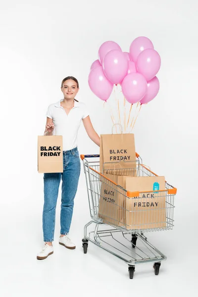 Jovem segurando saco de compras com letras sexta-feira preto perto do carrinho e balões rosa no branco — Fotografia de Stock