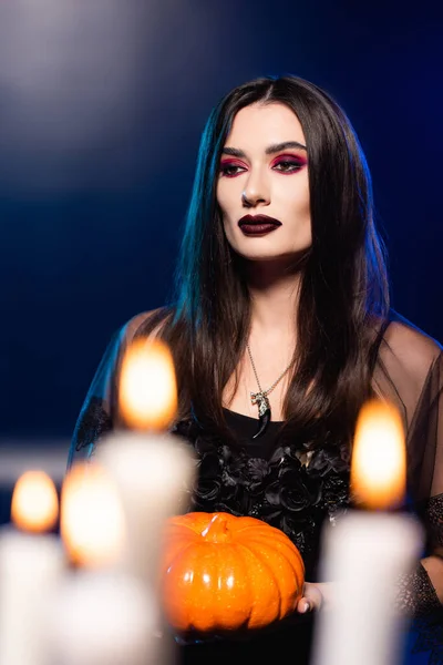 Selective focus of woman with black makeup holding pumpkin near burning candles on blue, halloween concept — Stock Photo