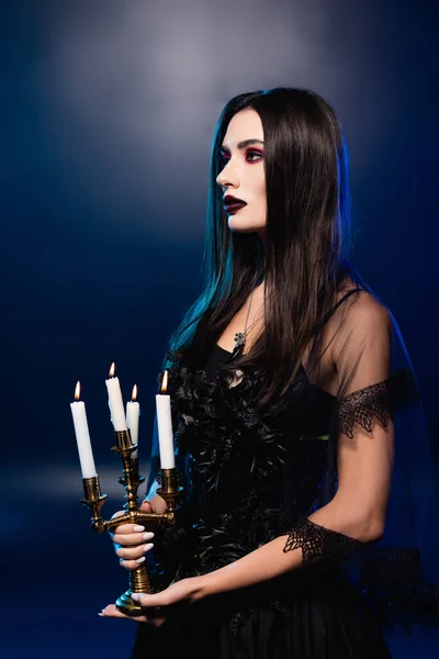 Mujer pálida con maquillaje negro sosteniendo candelabro con velas encendidas en azul, concepto de halloween - foto de stock