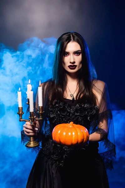 Молодая бледная женщина с черным макияжем держа тыкву и горящие свечи на голубом с дымом, Хэллоуин концепции — стоковое фото