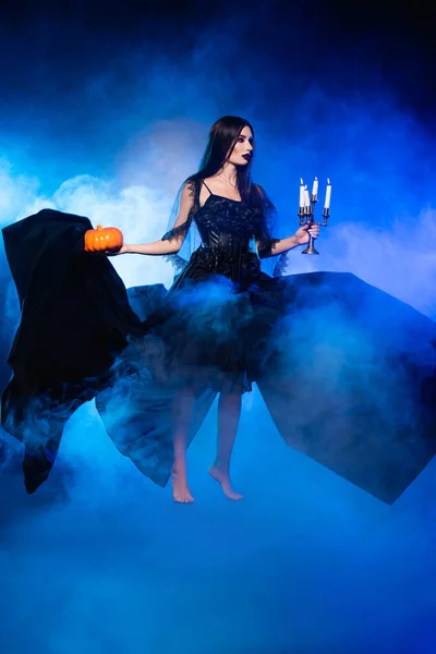 Young barefoot woman in black dress holding pumpkin and burning candles on blue with smoke — Stock Photo