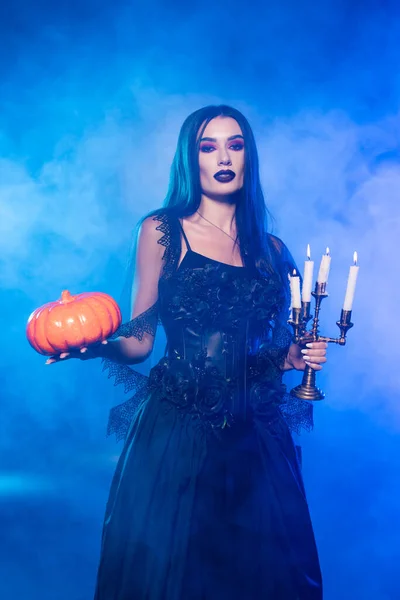 Joven mujer pálida en vestido negro sosteniendo calabaza y velas encendidas en azul con humo — Stock Photo