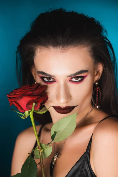 Молода жінка з темним макіяжем дивиться на камеру біля червоної троянди на синьому — стокове фото