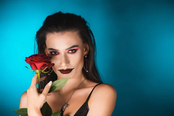 Mujer joven con maquillaje oscuro mirando a la cámara y sosteniendo rosa roja en azul - foto de stock