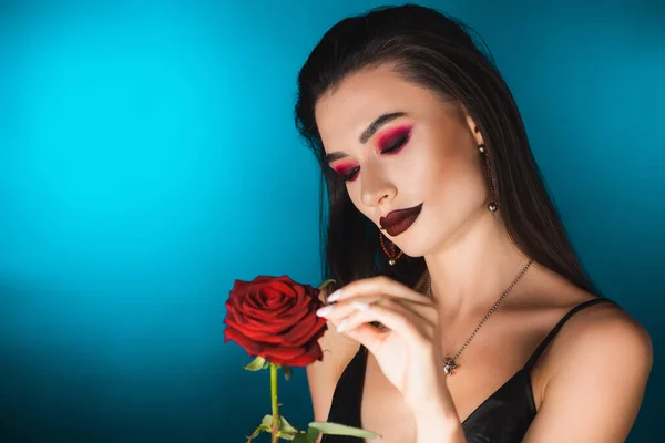 Молода жінка з темним макіяжем торкається червоної троянди на синьому — стокове фото
