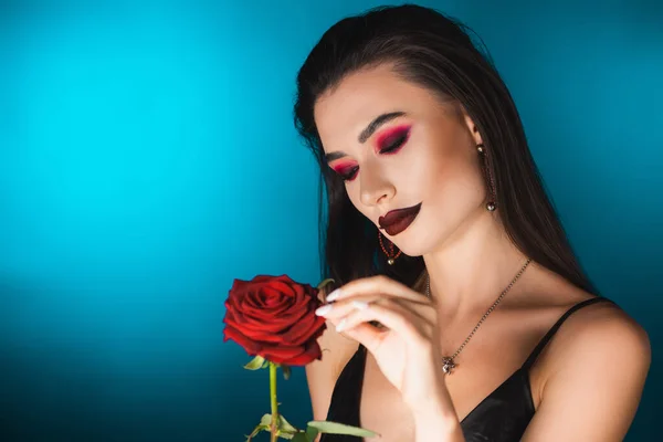 Mujer joven con maquillaje oscuro tocando rosa roja sobre azul - foto de stock