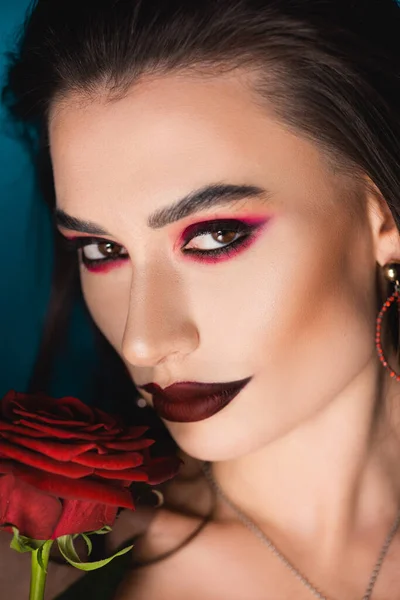 Крупним планом молода жінка з темним макіяжем біля червоної троянди на синьому — стокове фото