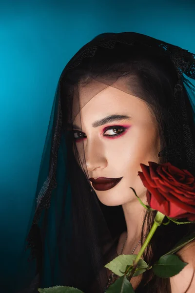 Mariée maléfique avec maquillage noir et voile regardant la caméra près de rose rouge sur bleu — Photo de stock