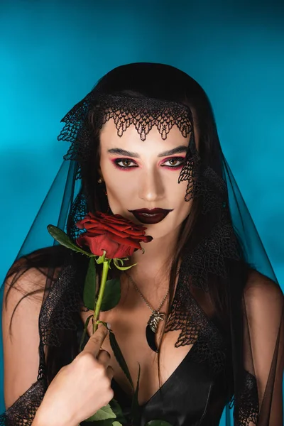 Зла жінка з чорним макіяжем і темною завісою тримає троянду на синьому — стокове фото