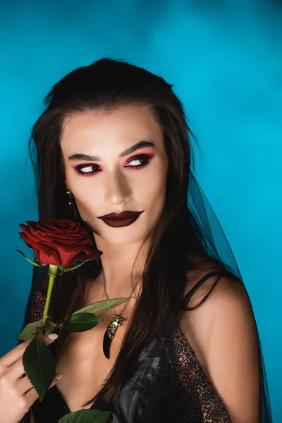 Geheimnisvolle junge Frau mit schwarzem Make-up im Schleier, die in der Nähe von Rose auf Blau wegschaut — Stockfoto