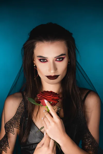 Misteriosa jovem com véu preto e maquiagem segurando rosa no azul — Fotografia de Stock