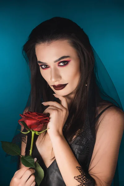 Mysterious young woman with black veil and makeup holding red rose on blue — Stock Photo