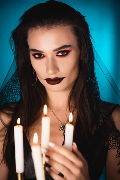 Selective focus of young woman with black makeup and veil near burning candles on blue — Stock Photo