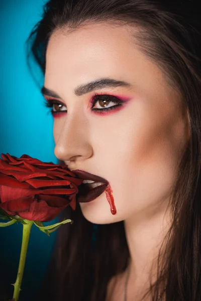 Mujer espeluznante joven con sangre en la cara cerca de rosa roja en azul - foto de stock