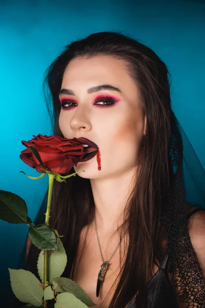 Mujer joven y espeluznante con sangre en la cara cerca de rosa roja en azul - foto de stock