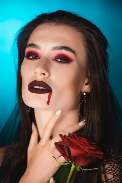 Jeune femme avec du sang sur le visage tenant rose rouge sur bleu — Photo de stock