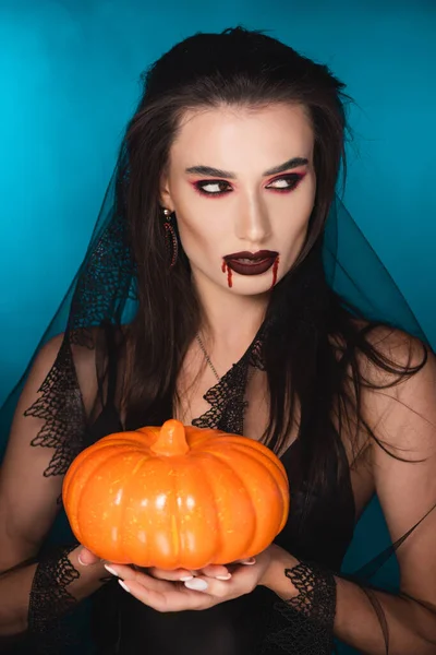 Woman in veil with red blood on face holding pumpkin on blue — Stock Photo