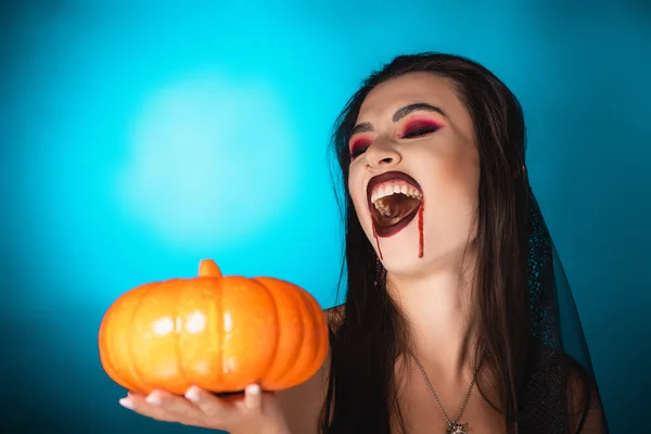 Femme effrayante avec du sang rouge sur le visage tenant citrouille et riant sur le bleu — Photo de stock